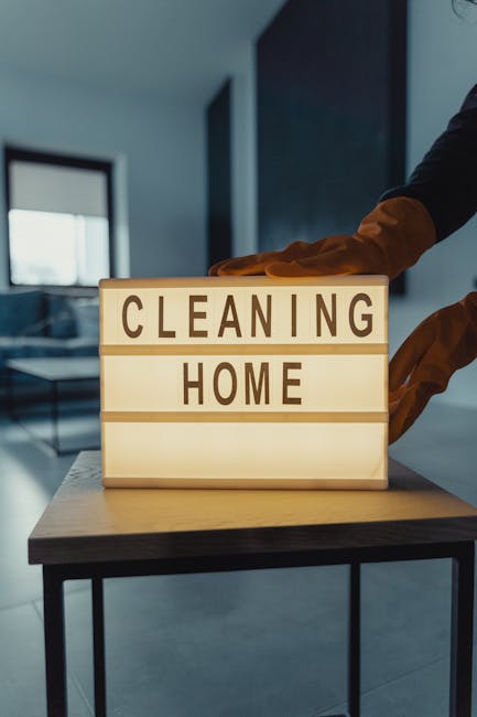 A person wearing black clothing and brown gloves is adjusting a lit-up sign with the words 'CLEANING HOME' in a modern, well-lit living room. The room features a dark sofa set, a glass coffee table, a