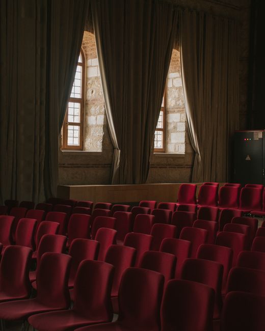 An indoor event space with a row of black plastic and chrome metal chairs arranged on a polished dark floor. The chairs are evenly spaced facing a stage area, which is partially obscured by closed bla